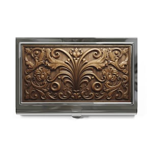 May include: A rectangular business card holder with a detailed, carved wooden panel featuring an ornate floral design. The wooden panel is set within a silver-colored metal frame. The design includes swirling leaves and symmetrical patterns.
