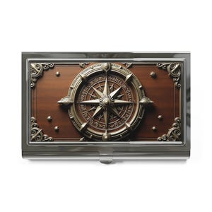 May include: A silver business card holder with a detailed compass design. The compass is bronze-colored and set against a dark wood background. The holder has ornate corner details and a push-button clasp.