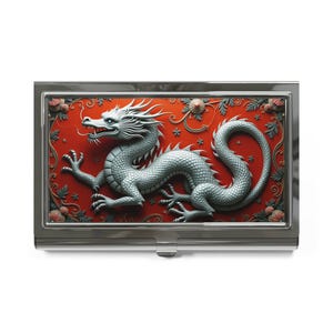 May include: A silver metal business card holder with a red background and a silver dragon design. The dragon is in a curled position with its head facing the left side of the image. The background is decorated with floral patterns and stars.