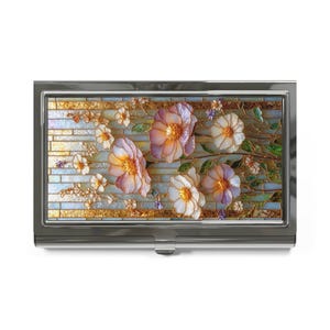 May include: A silver metal business card holder with a floral design. The design features pink and white flowers with green leaves on a background of iridescent tiles.