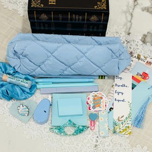 May include: A light blue quilted pencil case with a variety of school supplies, including pens, pencils, a lip balm, a bookmark, and a small blue pouch. The supplies are arranged on a white lace surface.