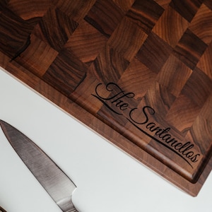 May include: A dark brown end-grain cutting board with a rectangular shape and a juice groove. The board has a checkerboard pattern and the engraved text "The Santanellos". A stainless steel knife is next to the board.