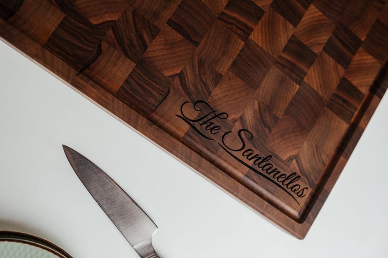 May include: A dark brown end-grain cutting board with a rectangular shape and a juice groove. The board has a checkerboard pattern and the engraved text "The Santanellos". A stainless steel knife is next to the board.