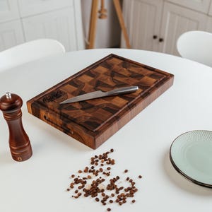 May include: A rectangular wooden cutting board with a checkerboard pattern, a stainless steel knife, and a wooden pepper grinder. The cutting board has the text "Carrie's" engraved on it. Scattered peppercorns and a plate are also on the white surface.