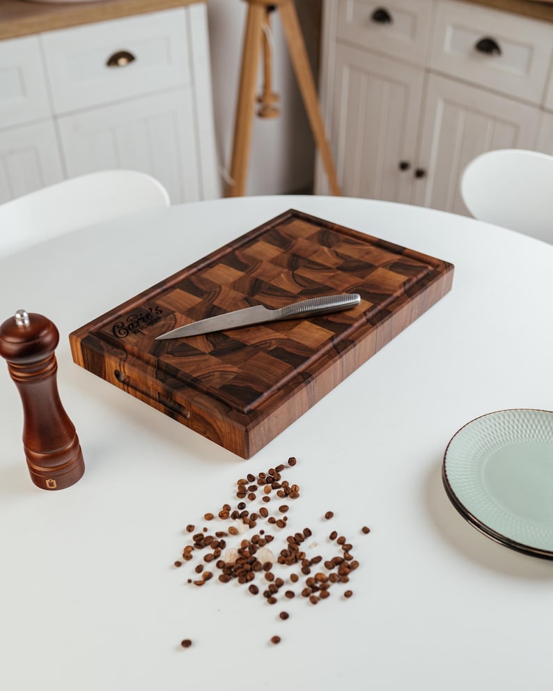 May include: A rectangular wooden cutting board with a checkerboard pattern, a stainless steel knife, and a wooden pepper grinder. The cutting board has the text "Carrie's" engraved on it. Scattered peppercorns and a plate are also on the white surface.