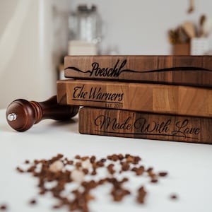 May include: A stack of three wooden cutting boards with engraved text. The top board reads "Poeschl", the middle board says "The Warners EST. 2019", and the bottom board says "Made With Love". A wooden pepper grinder and peppercorns are in the foreground.