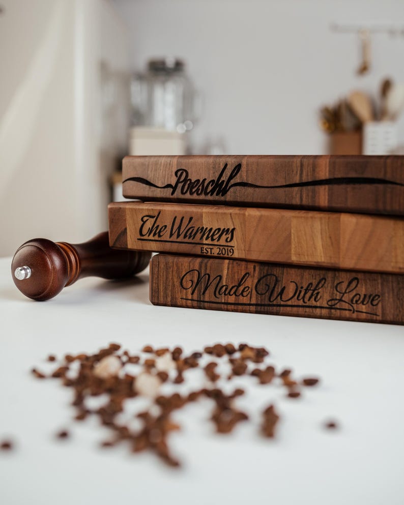 May include: A stack of three wooden cutting boards with engraved text. The top board reads "Poeschl", the middle board says "The Warners EST. 2019", and the bottom board says "Made With Love". A wooden pepper grinder and peppercorns are in the foreground.