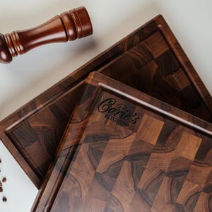 May include: Two dark brown wooden cutting boards with a checkerboard pattern and a raised edge. A wooden pepper grinder rests above the boards. The text "Carie's Kitchen" is visible on one board. Coffee beans are scattered around the boards.