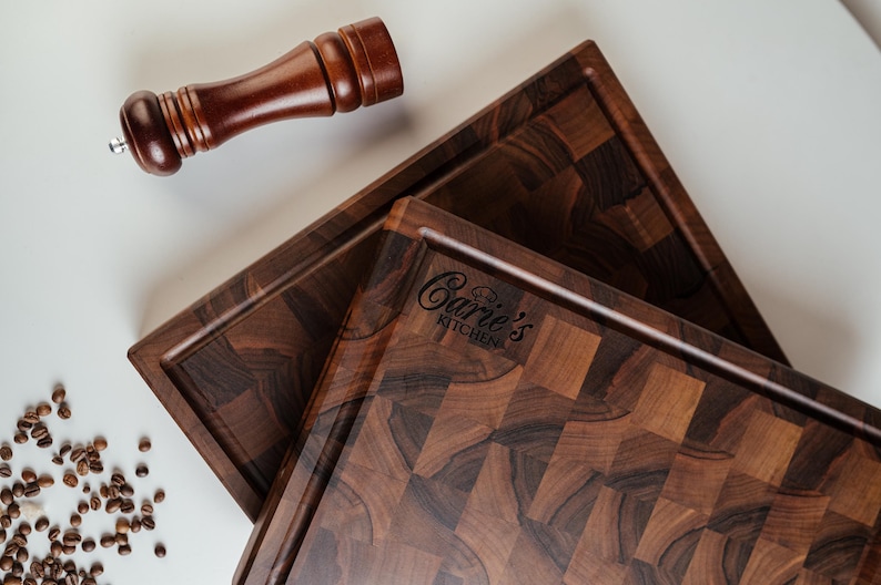 May include: Two dark brown wooden cutting boards with a checkerboard pattern and a raised edge. A wooden pepper grinder rests above the boards. The text "Carie's Kitchen" is visible on one board. Coffee beans are scattered around the boards.