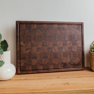May include: A rectangular end-grain chopping board with a dark brown, chequered pattern. The board has a beveled edge and the name "The Santanellos" is engraved on the lower right. The board is displayed on a wooden surface.