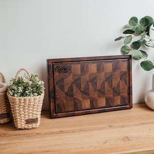 May include: A dark brown wooden cutting board with a checkered pattern and a carved border. The board has the text "Care's Kitchen" in the upper right corner. Two small woven baskets with plants and a white vase with greenery are also visible.