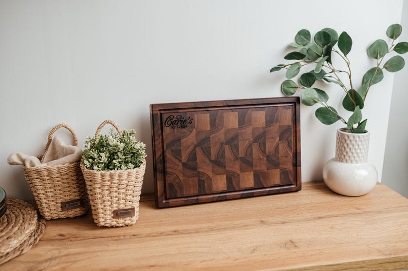 May include: A dark brown wooden cutting board with a checkered pattern and a carved border. The board has the text "Care's Kitchen" in the upper right corner. Two small woven baskets with plants and a white vase with greenery are also visible.