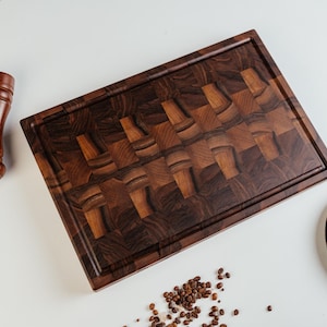 May include: A rectangular, dark brown wooden cutting board with a patterned design. A wooden pepper grinder, a stainless steel knife, and a small plate are also present. Scattered coffee beans add a decorative touch. The items are arranged on a white surface.