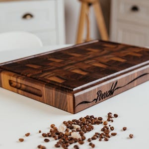 May include: A rectangular, dark brown wooden cutting board with a patterned surface and a carved handle. The word "Poeschl" is engraved on the side. Coffee beans and sugar crystals are scattered on the white surface.