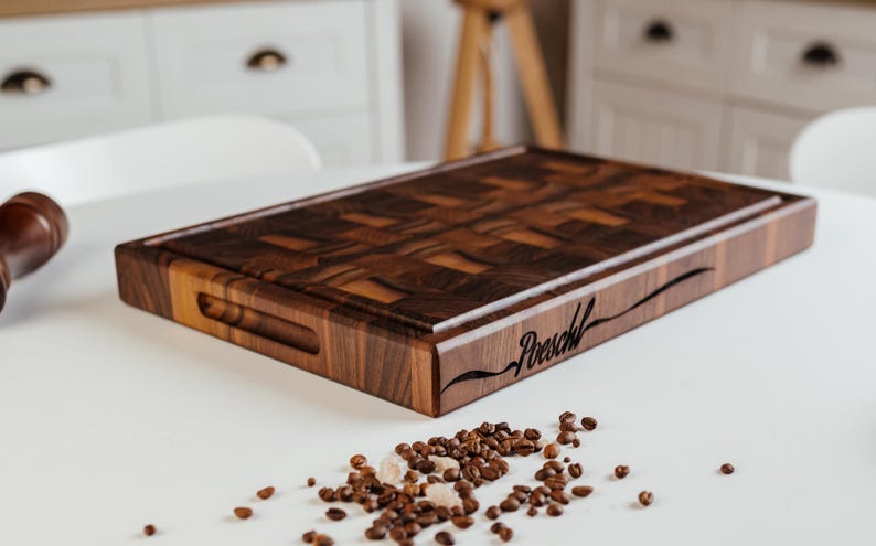 May include: A rectangular, dark brown wooden cutting board with a patterned surface and a carved handle. The word "Poeschl" is engraved on the side. Coffee beans and sugar crystals are scattered on the white surface.