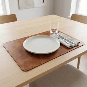Op de afbeelding: Een rechthoekige bruine placemat van kunstleer met gestikte randen, met daarop een wit gespikkeld keramieken bord, een helder glazen beker, een zilveren vork en een zilveren mes op een gevouwen lichtgrijze linnen servet.