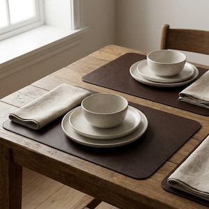 May include: A dining table setting featuring dark brown rectangular placemats, beige linen napkins, and off-white speckled ceramic dinnerware sets. Each set includes a round dinner plate, a smaller salad plate, and a deep cereal bowl.