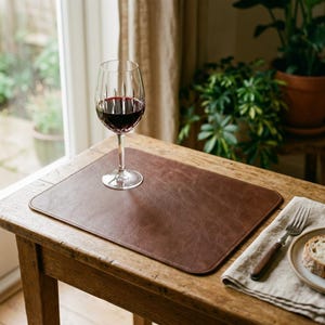 May include: A rectangular brown leather placemat with rounded corners, featuring a clear glass wine goblet filled with red wine, a silver fork with a wooden handle, and a slice of bread on a beige linen napkin.