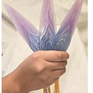 May include: A cluster of three stylized, petal-shaped objects in shades of blue and purple, held together with a gold tassel. The objects have a textured surface with a decorative pattern. The overall design suggests a fantasy or decorative theme.