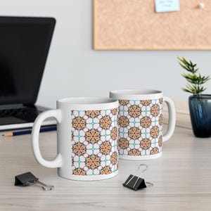 May include: Two white ceramic mugs with a repeating floral pattern in orange, yellow, red, and teal. The mugs are on a light-colored wooden surface, with a laptop and office supplies in the background.