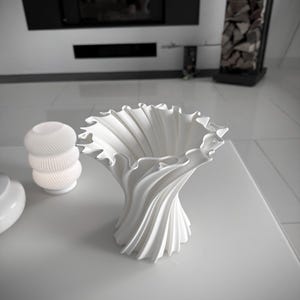 May include: A white, sculptural vase with a swirling, ruffled design. The vase has a wide, open top and a narrow base. The design is reminiscent of fabric folds. The vase is sitting on a white surface.