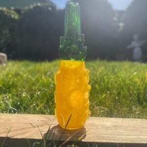 Puede incluir: Una pipa de vidrio amarilla y verde con forma de piña. La pipa tiene un pequeño agujero en el lateral y una parte superior verde.
