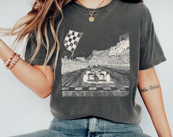 May include: A charcoal grey short-sleeve t-shirt featuring a white graphic print of a race car on a track, a chequered flag, and a grandstand filled with spectators. The design depicts a high-speed racing scene with detailed line work.