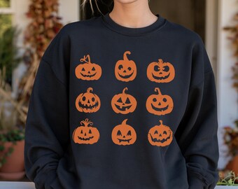 May include: A black long-sleeve sweatshirt featuring a graphic print of nine orange jack-o'-lanterns arranged in a three-by-three grid. Each pumpkin displays a unique carved face with a distressed texture, set against the solid black fabric.