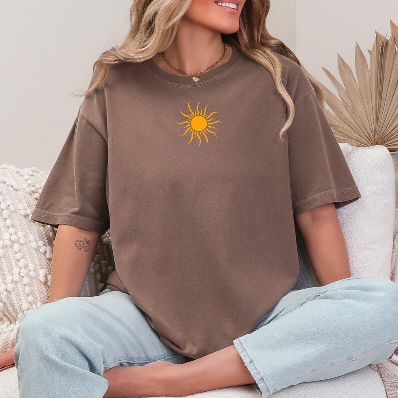 May include: A brown t-shirt with a yellow sun graphic. The short-sleeved shirt is a solid color and has a relaxed fit. The person is wearing light blue jeans.