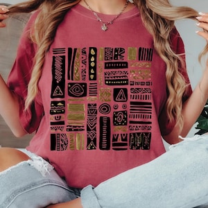 May include: A maroon t-shirt featuring a geometric design in black and gold. The design is composed of various shapes and patterns arranged in a grid-like structure. The shirt is a solid maroon color.