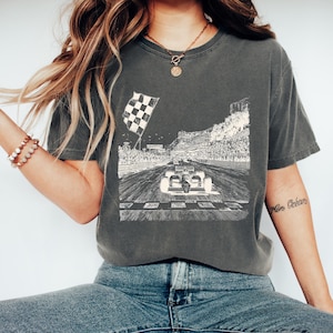 May include: Dark grey t-shirt with a black and white illustration of a race car approaching a chequered flag. The design includes a racetrack, grandstands, and spectators. The shirt is a casual style.