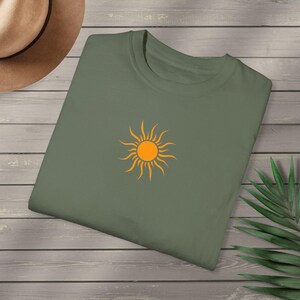 May include: Olive green t-shirt featuring a bright orange sun graphic. The sun design is centered on the front of the shirt. The shirt is folded and placed on a wooden surface, with a hat visible in the background.
