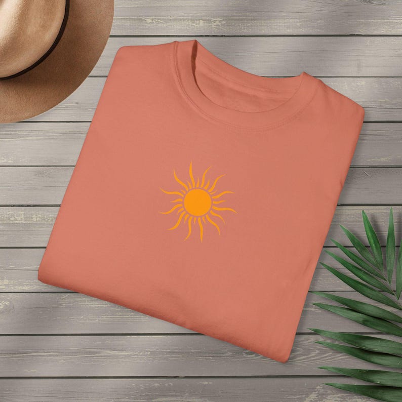 May include: A coral-colored t-shirt featuring a bright orange sun graphic. The sun design is centered on the front of the shirt. The shirt is folded neatly on a wooden surface, with a hat and a green plant visible in the background.