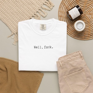 May include: A white t-shirt with the text "Well, fuck." is displayed alongside tan pants, a woven placemat, and a candle. The shirt is made of a soft material and is likely for casual wear.