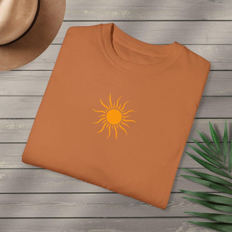 May include: An orange t-shirt with a round neck and short sleeves. The shirt features a bright orange sun graphic in the center. The shirt is folded and placed on a wooden surface, with a hat and a plant in the background.