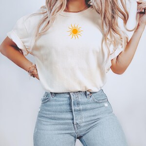 May include: Cream-colored t-shirt featuring a yellow sun graphic on the front. The shirt has short sleeves and is paired with light blue denim jeans. The overall style is casual and relaxed.