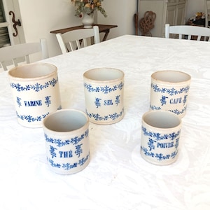 Peut inclure: Cinq pots en céramique blanc cassé avec des motifs floraux bleus et des étiquettes. Les étiquettes indiquent "FARINE", "SEL", "CAFE", "THE" et "POIVRE". Ces récipients vintage sont pour la cuisine.