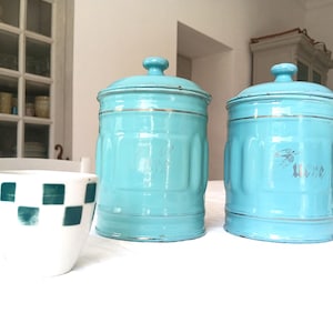 Peut inclure: Deux boîtes de conservation cylindriques en métal turquoise avec couvercles assortis et bordures argentées, placées à côté d'une tasse en céramique blanche à motif à carreaux verts. Les boîtes présentent des nervures verticales et du texte.