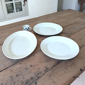 Peut inclure: Deux assiettes rondes en céramique blanche, un plat ovale en céramique blanche et une petite tasse à expresso blanche ornée d'un motif à carreaux verts et blancs sur l'extérieur. Les objets sont disposés sur une surface en bois.