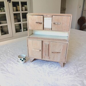 Peut inclure: Un petit buffet en bois clair avec deux portes en bas et deux portes plus petites en haut. Le buffet a un design simple et est posé sur une nappe blanche. Une petite tasse à thé à carreaux est devant le buffet.
