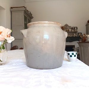 Peut inclure: Un grand pot en grès cylindrique doté d'une glaçure lisse beige clair aux nuances grisâtres subtiles. Le récipient présente un bord épais et large ainsi que deux petites anses intégrées sur les côtés. La surface montre des variations naturelles.