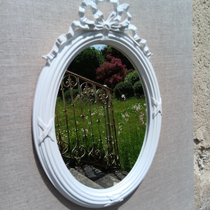 Peut inclure: Miroir ovale blanc avec un nœud décoratif en haut. Le miroir reflète une scène de jardin avec une clôture en fer forgé, de l'herbe verte et des arbres. Le cadre a un design texturé et nervuré.