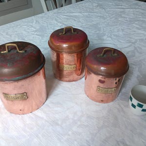 Peut inclure: Trois boîtes en cuivre avec couvercles et poignées en laiton, étiquetées "SUCRE", "CAFÉ" et "THÉ". Une petite tasse blanche avec un motif à carreaux verts est à proximité. Les boîtes ont un aspect vintage et vieilli.