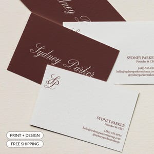 May include: Business cards with the name "Sydney Parker" printed in elegant script. The cards are in shades of white and burgundy, with the company's contact information and logo. The words "Print + Design" and "Free Shipping" are also visible.