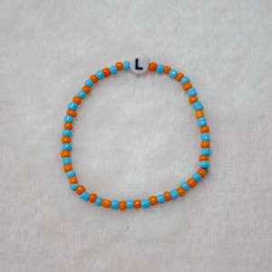 May include: A blue and orange beaded bracelet with a white bead with the letter "L" on it.