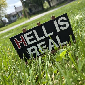 May include: A black sign with the words "HELL IS REAL" in white and red lettering, displayed on a grassy lawn. The sign is held up by small wooden supports, with a blurred background of a residential area.