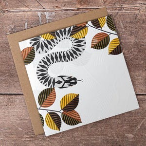 May include: A square greeting card featuring a stylised black and white patterned snake winding through branches with yellow and copper-coloured leaves. The white card surface has a subtle embossed leaf texture and sits atop a brown kraft paper envelope.