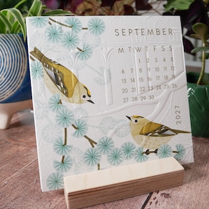 May include: A square 2027 calendar page for September featuring two gold and brown birds perched on branches with teal circular floral patterns. The white paper has an embossed architectural arch design and is held by a light wood block stand.