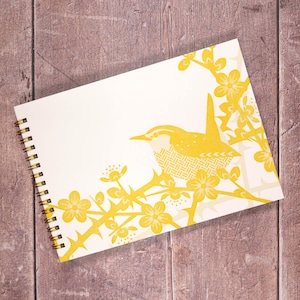 May include: A cream-coloured, spiral-bound notebook with a yellow illustration of a bird on a flowering branch. The bird and floral design are in a graphic style. The notebook is on a wooden surface.