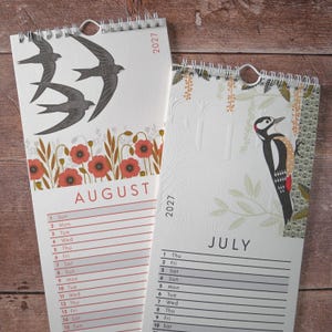 May include: Two 2027 wall calendars with spiral binding. The August page features three black swallows flying above red poppies. The July page displays a woodpecker perched on a tree branch with green leaves and a textured white background.